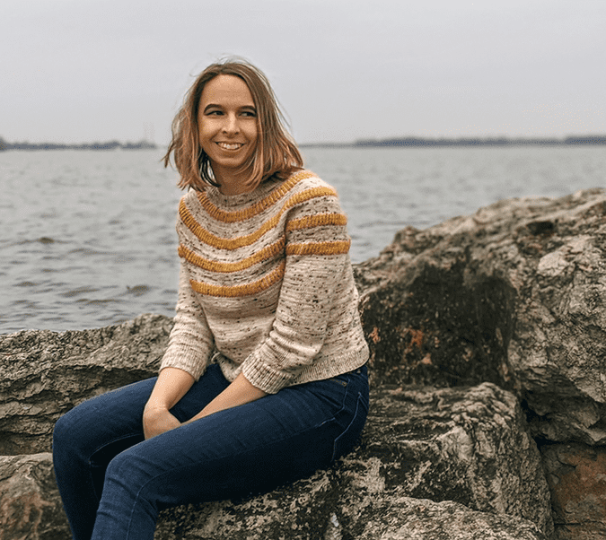 Rachel sitting on a boulder in front of Lake Erie, wearing a sweater and jeans.